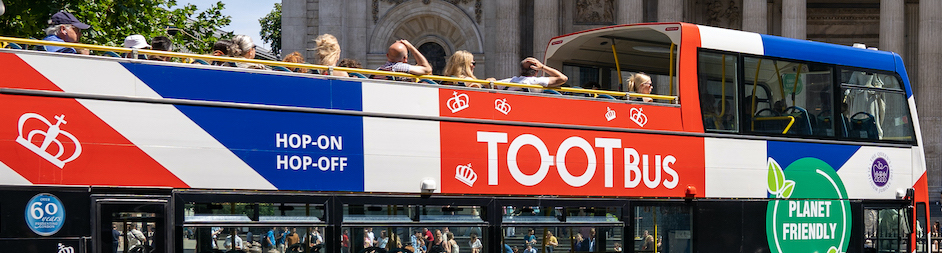 Group of travellers taking selfies on a Tootbus London open-top sightseeing bus with the River Thames and city skyline in the background.