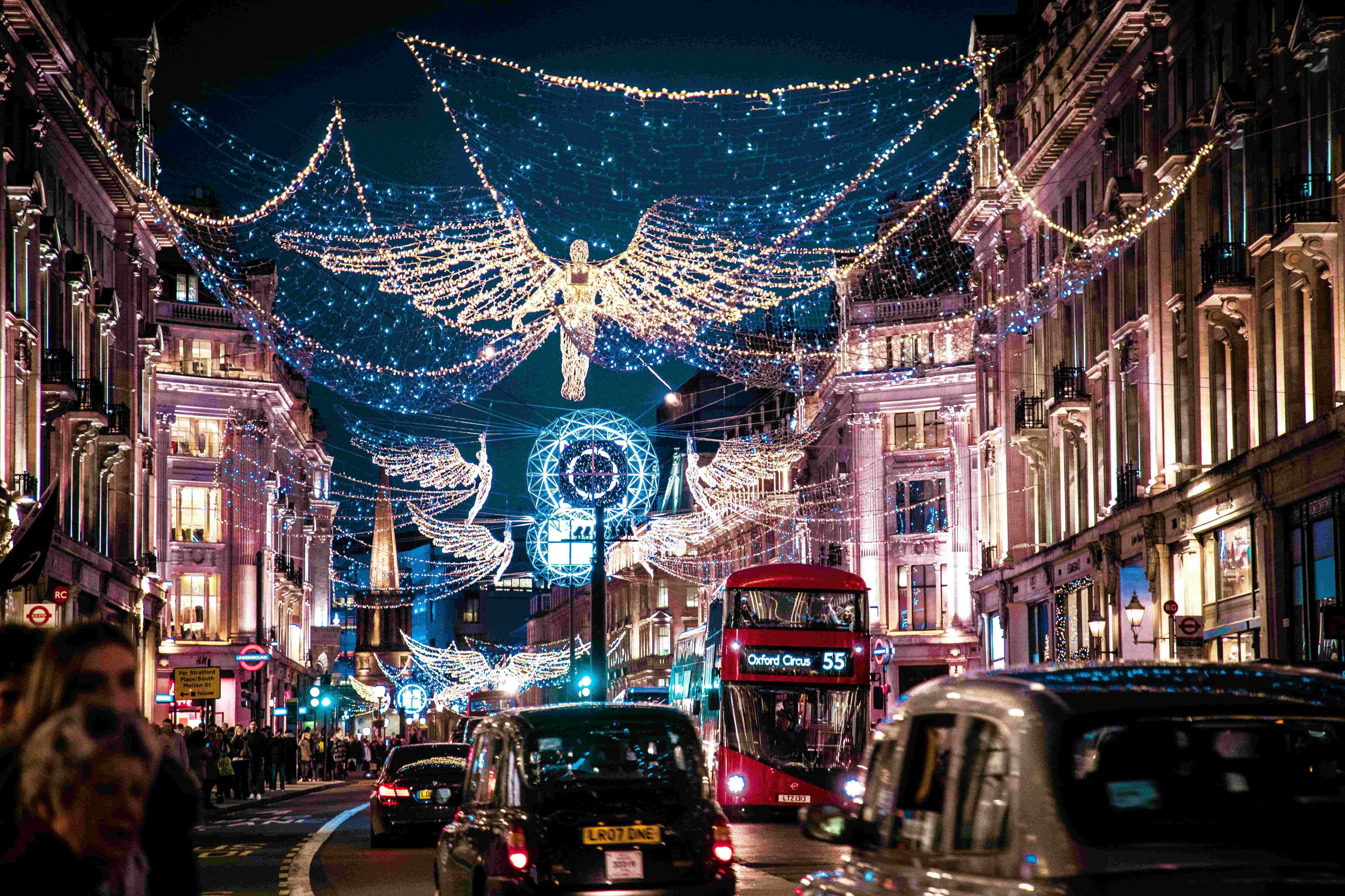 Oxford Street in London illuminated by dazzling Christmas angel lights strung above the bustling road. A red double-decker bus heading to Oxford Circus and classic black London taxis are surrounded by shoppers enjoying the festive atmosphere.