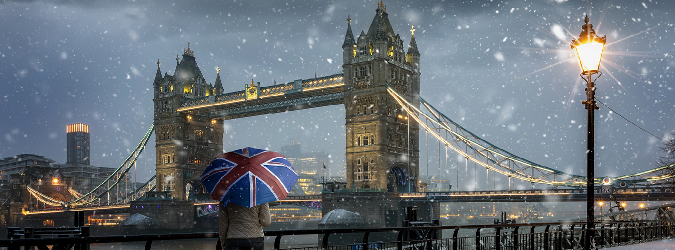 A young couple stands side-by-side on a bridge, gazing out at the iconic London skyline. Big Ben and the Houses of Parliament are visible in the distance